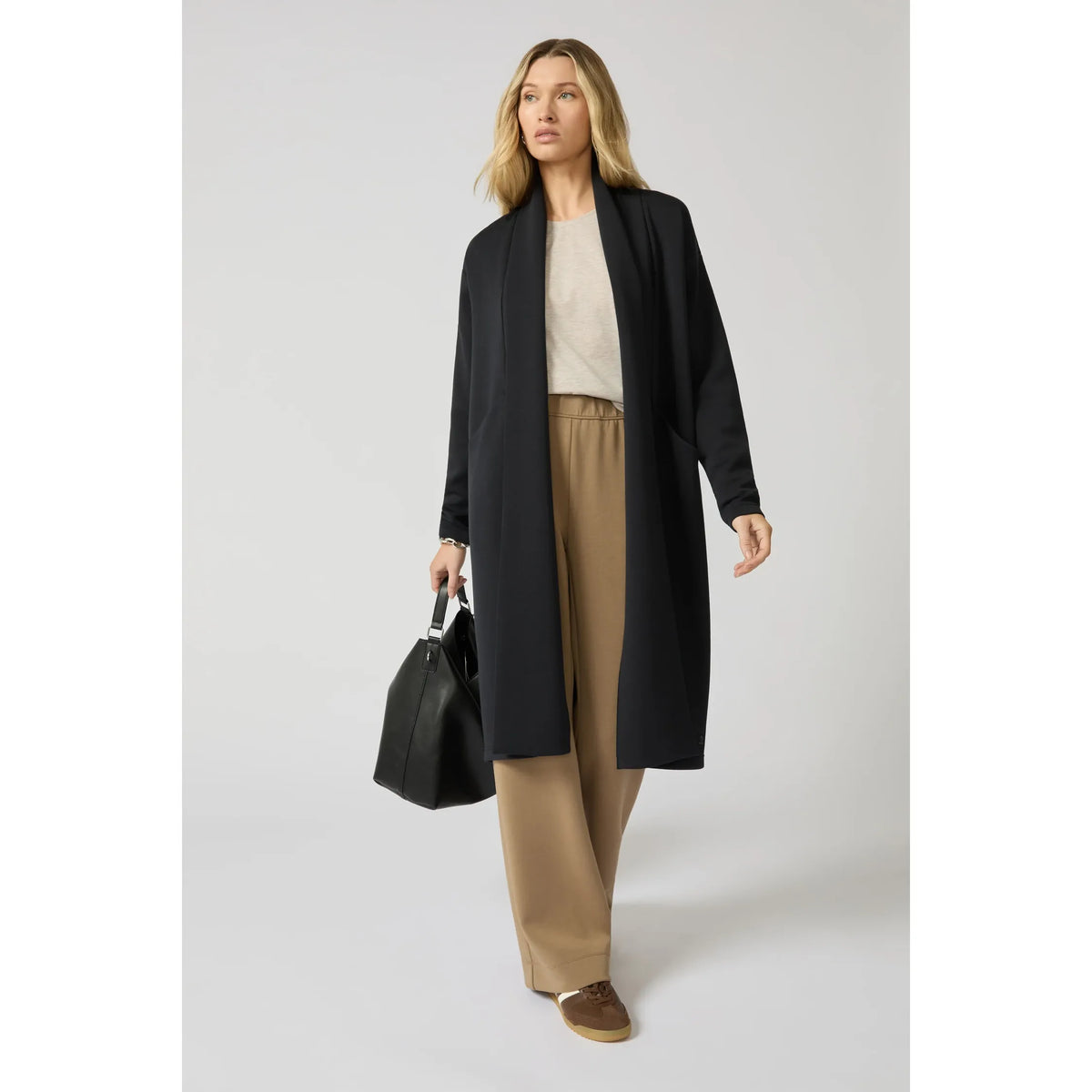 Model wearing a black long cardigan with a clean longline silhouette, styled with tan wide-leg trousers and a black handbag.