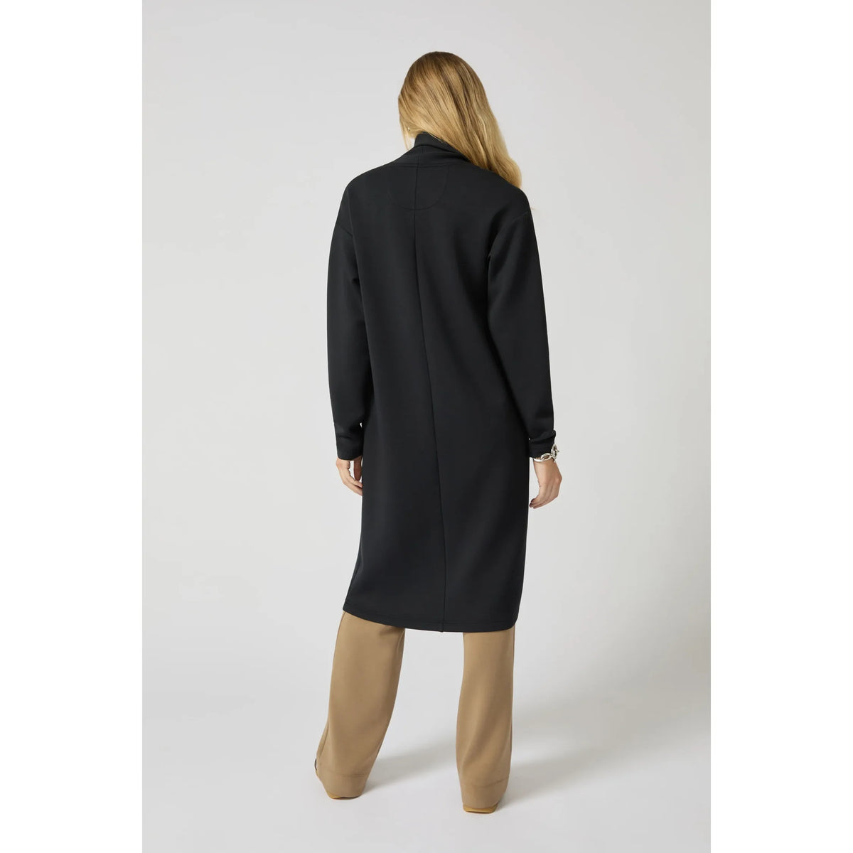 Back view of a black long cardigan with a clean longline silhouette and center seam, worn with tan trousers.