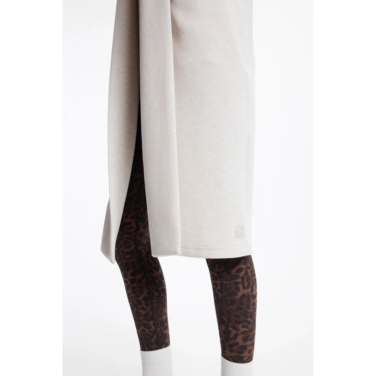 Light beige long cardigan with high side slit and small logo patch, styled over leopard print leggings and white socks.