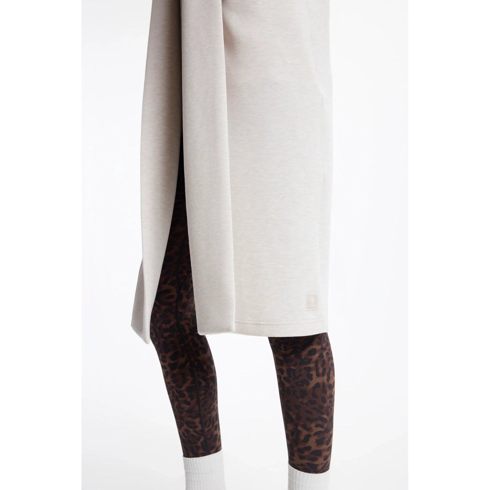 Light beige long cardigan with high side slit and small logo patch, styled over leopard print leggings and white socks.