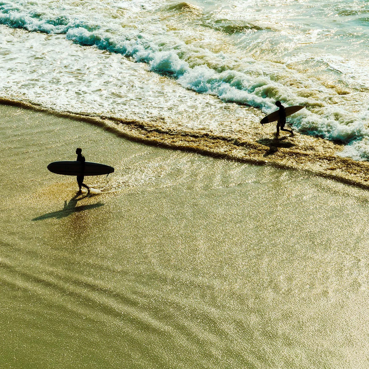 Two surfers walk along a sunlit beach with surfboards as waves crash onto the shore.