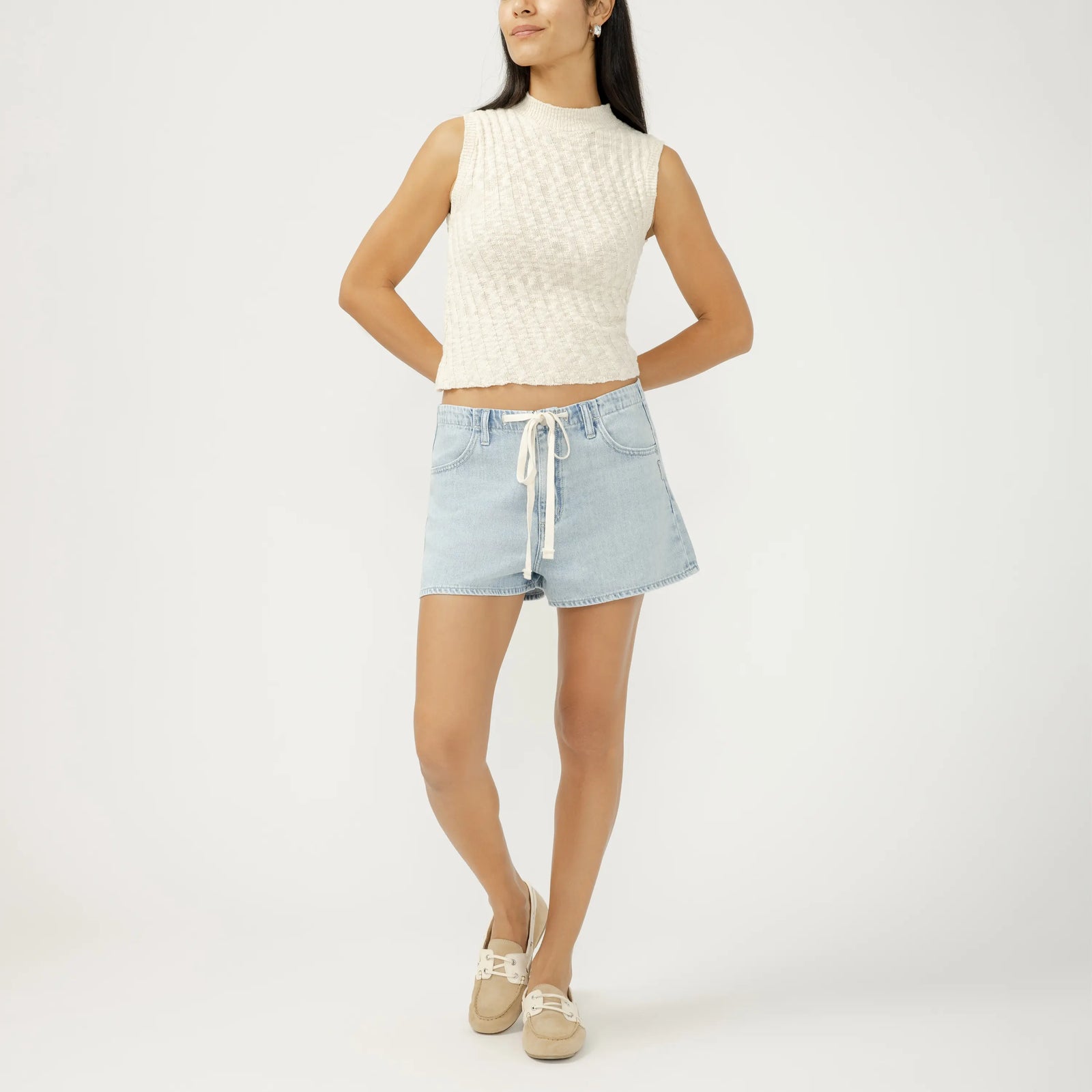 Model wearing a light denim jacket, beige ribbed top, and sage green drawstring shorts with white sneakers.