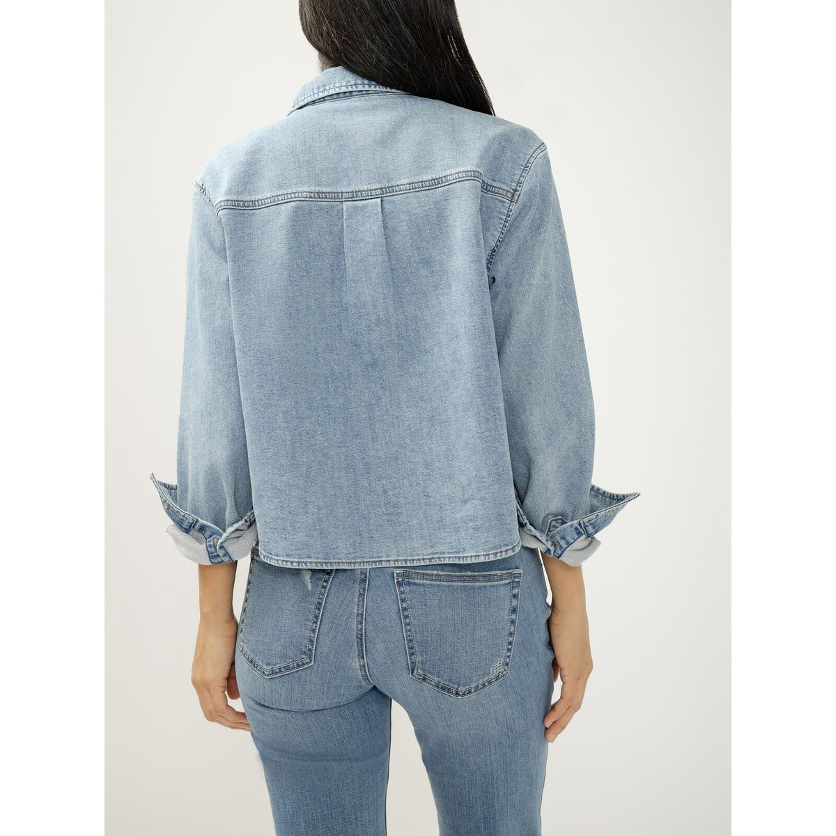 Back view of a woman wearing a light wash Silver Jeans button front knit flex shirt with rolled sleeves, paired with matching denim jeans.