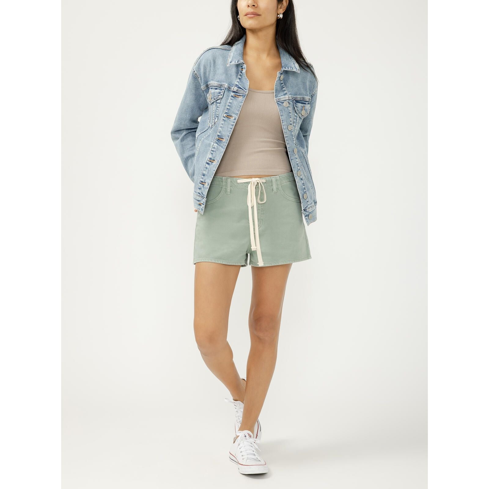 Model wearing a light denim jacket, beige ribbed top, and sage green drawstring shorts with white sneakers.