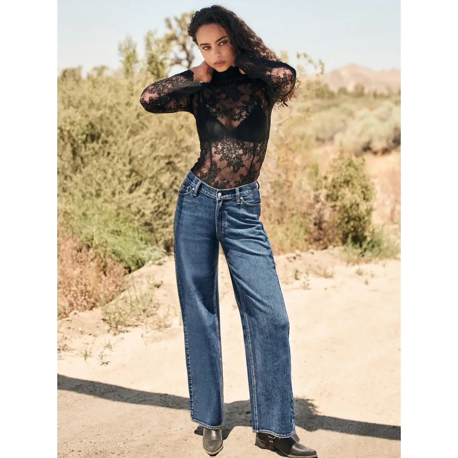 Model wearing silver V front wide leg jeans in root wash with a relaxed fit, paired with a black lace top in a desert setting.