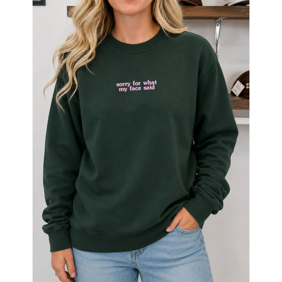 Dark green crew neck sweatshirt featuring pink embroidered text "sorry for what my face said" on the chest.