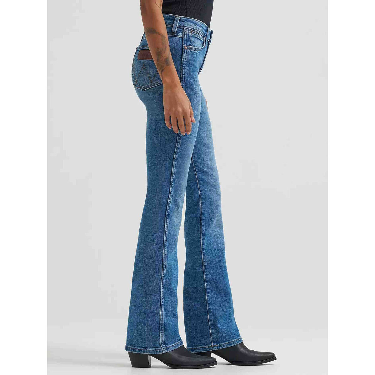 Side view of a woman wearing Wrangler Retro Bailey High Rise Bootcut Jeans with a subtle 17-inch flare over boots.