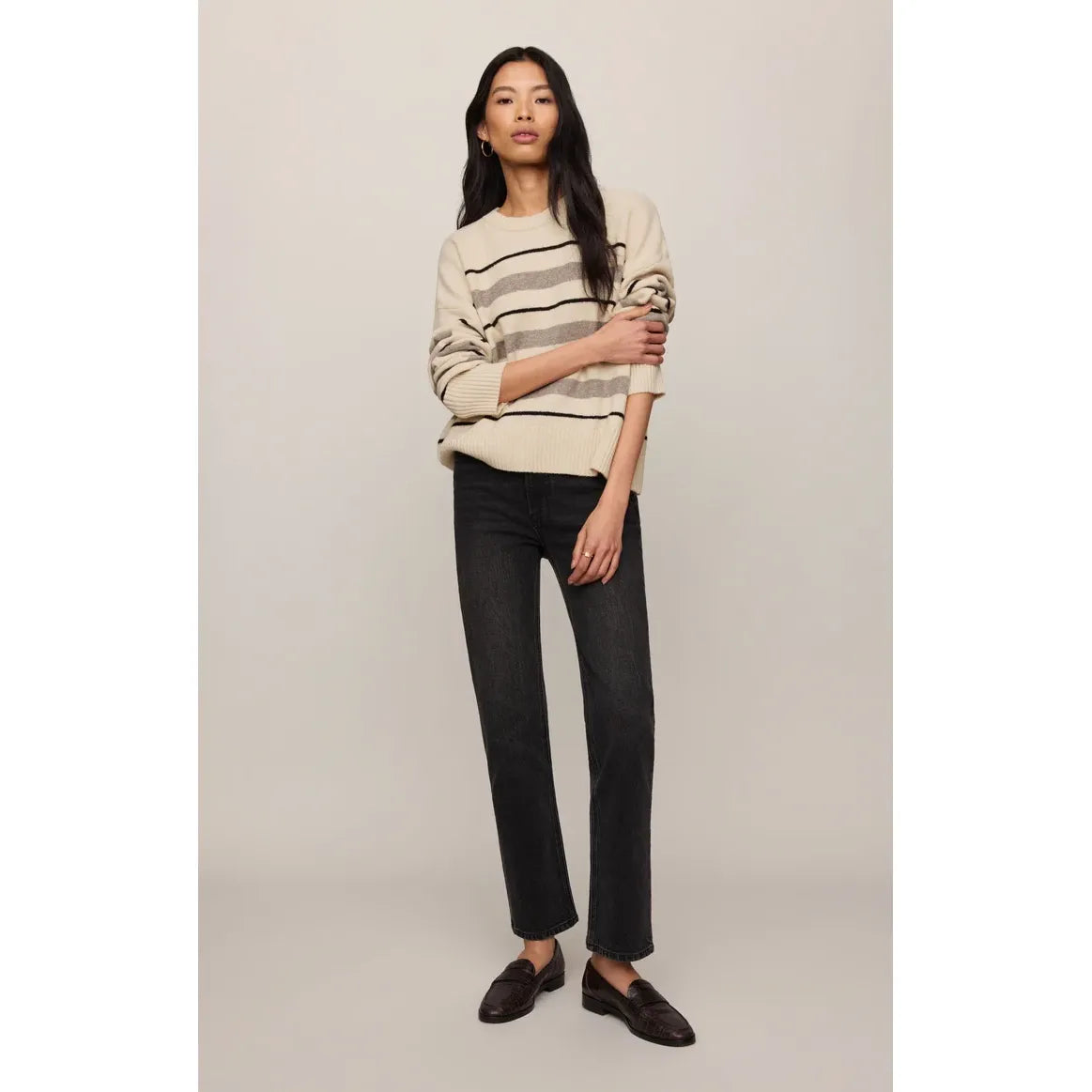 Model wears a cream Z Supply Cirrus striped sweater with black and grey horizontal stripes, paired with dark straight-leg jeans and brown loafers.