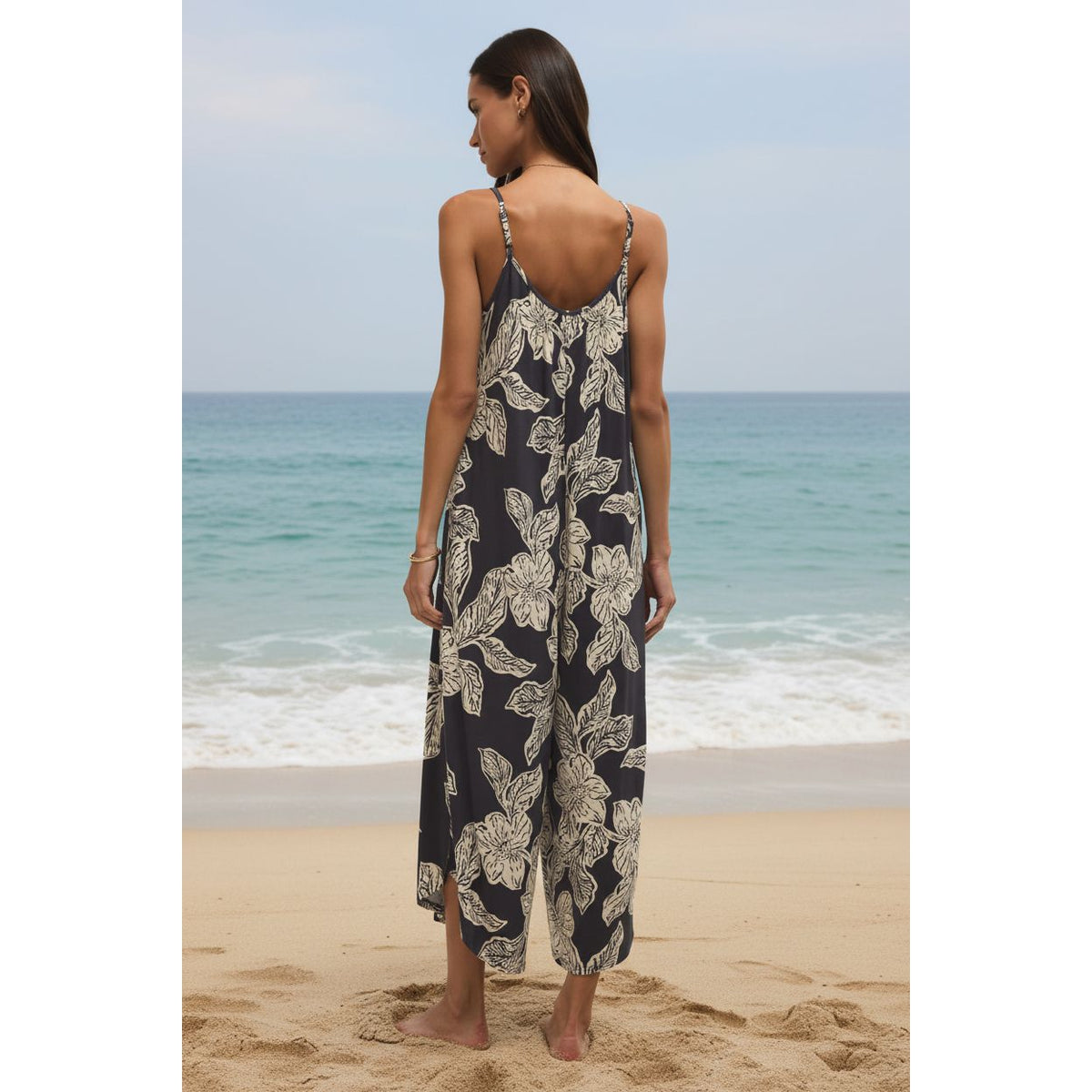 Back view of a model wearing Z Supply Flared Asha Floral Jumpsuit in navy with white florals on gauze at the beach.