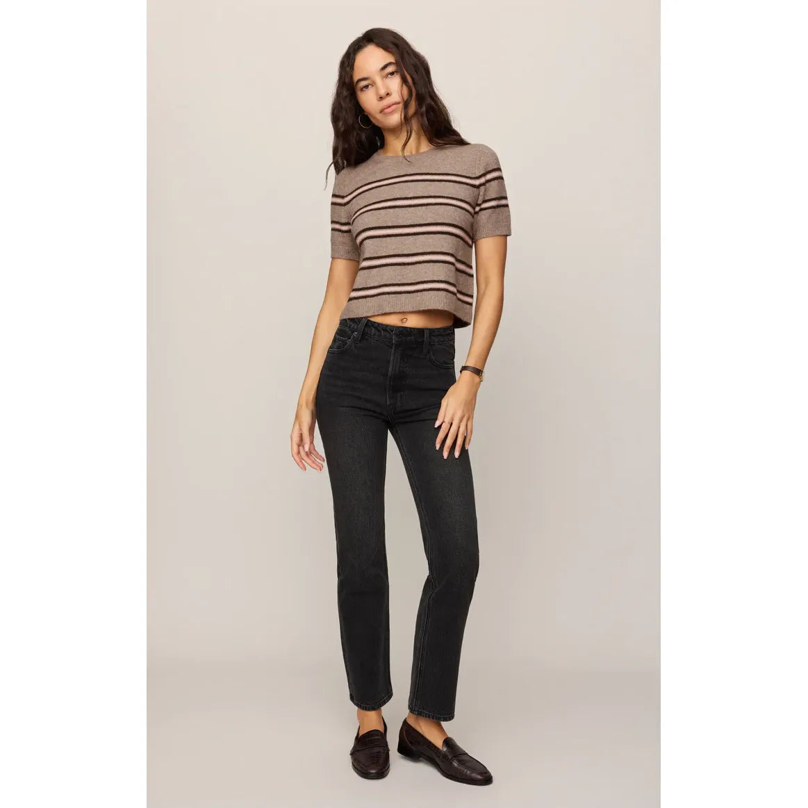 Woman wearing a cropped striped sweater tee in taupe with dark horizontal stripes, paired with black straight-leg jeans and brown loafers.