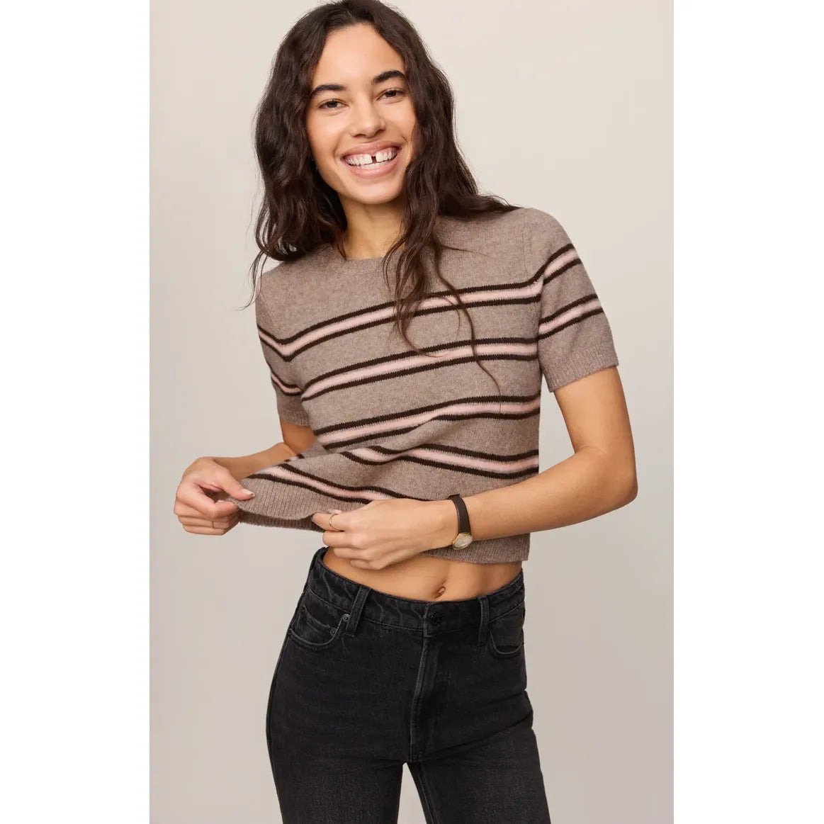 Woman smiling in a taupe short-sleeve striped sweater tee with pink and dark brown horizontal stripes, paired with black jeans.